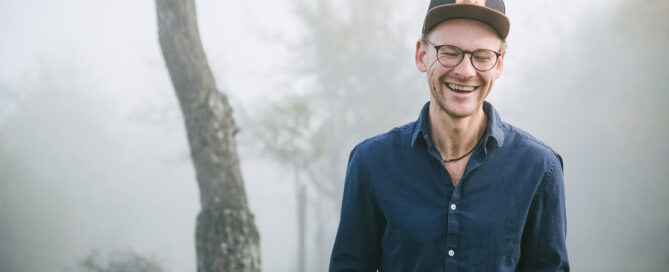 Christian Bekmulin - Musikerporträt für Website und Presse,  mit Brille und schwarzer Kappe steht Christian Bekmulin lächelnd im nebligen Park auf grünem Gras.