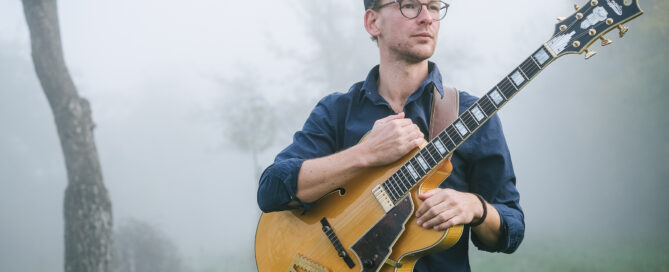 Christian Bekmulin mit Jazzgitarre steht im nebligen Freien vor Bäumen und Gras.