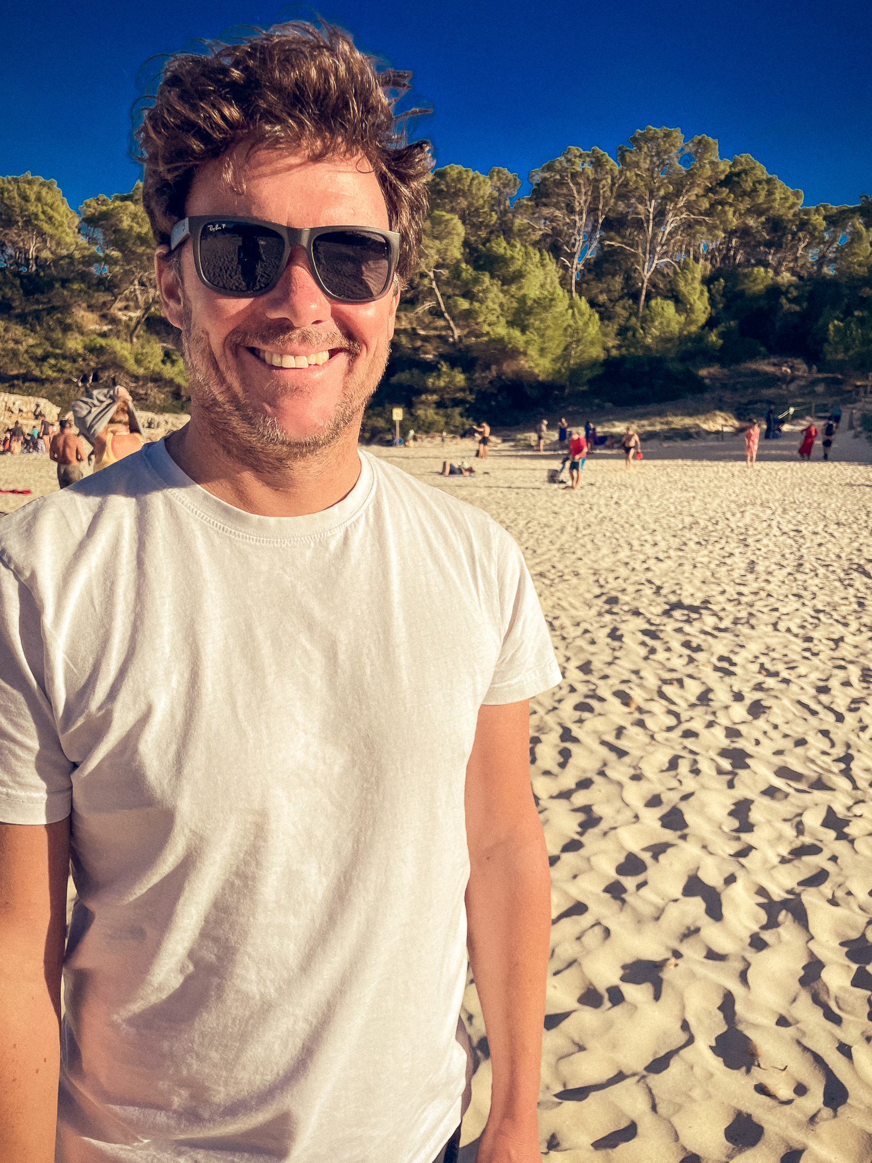 Entspannter Mann am Strand bei Sonnenuntergang Mann lächelnd am Strand bei Sonnenuntergang, trägt weißes T-Shirt, trägt Sonnenbrille, umgeben von Natur.