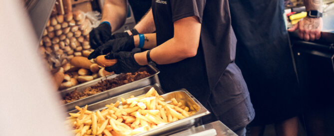 Drei Küchenmitarbeiter in schwarzer Uniform bereiten in einer Imbissküche frisch frittierte Pommes zu.