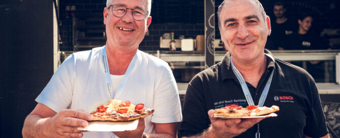 Zwei Männer halten Teller mit Pizza vor einem Foodtruck bei einem Außenevent.