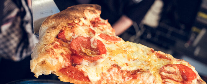 Frisch gebackene Pepperoni-Pizza wird mit Spatel serviert, unscharfe Personen im Hintergrund.