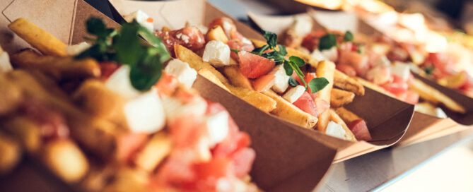Pommes frites mit Feta, Tomatenstücken und frischen Kräutern in braunen Papptabletts serviert.