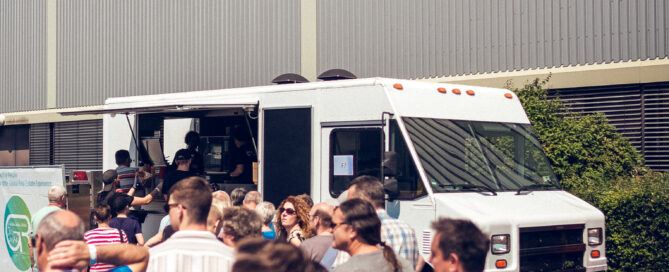 Menschen stehen bei sonnigem Wetter an einem weißen Foodtruck vor einer Industriehalle an.