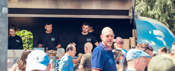 Menschen stehen an einem blauen Foodtruck im Freien und warten auf ihre Bestellungen.