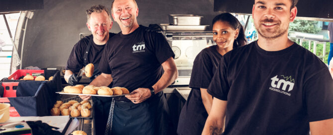 Vier Mitarbeitende in schwarzer Kleidung bereiten in einem Foodtruck Brotbrötchen auf einem Tablett vor.