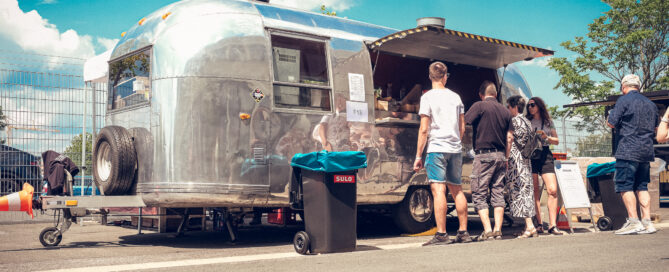 Menschen stehen vor einem silbernen Foodtruck mit Markise unter blauem Himmel an.