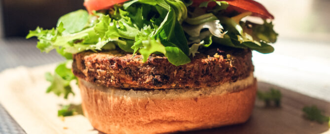 Veggie-Burger auf brauner Paperschale bei Tageslicht, aufgenommen vor unscharfem metallischem Hintergrund.