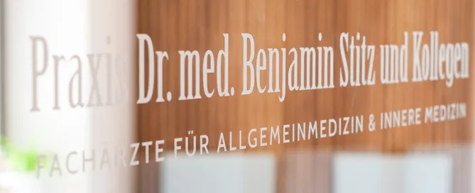 Glaswand einer Arztpraxis mit Aufschrift „Praxis Dr. med. Benjamin Stitz und Kollegen“ und Familienpiktogrammen.