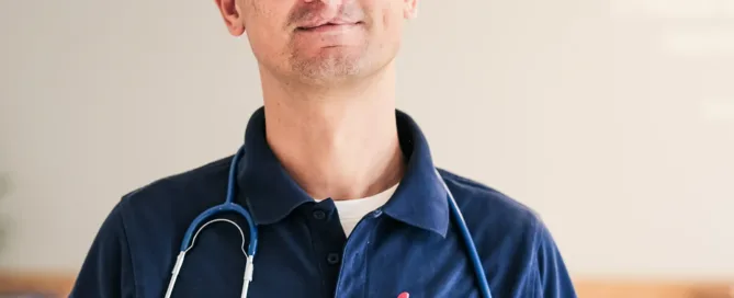 Arzt mit Stethoskop in blauer Kleidung steht in heller Praxis vor neutralem Hintergrund.