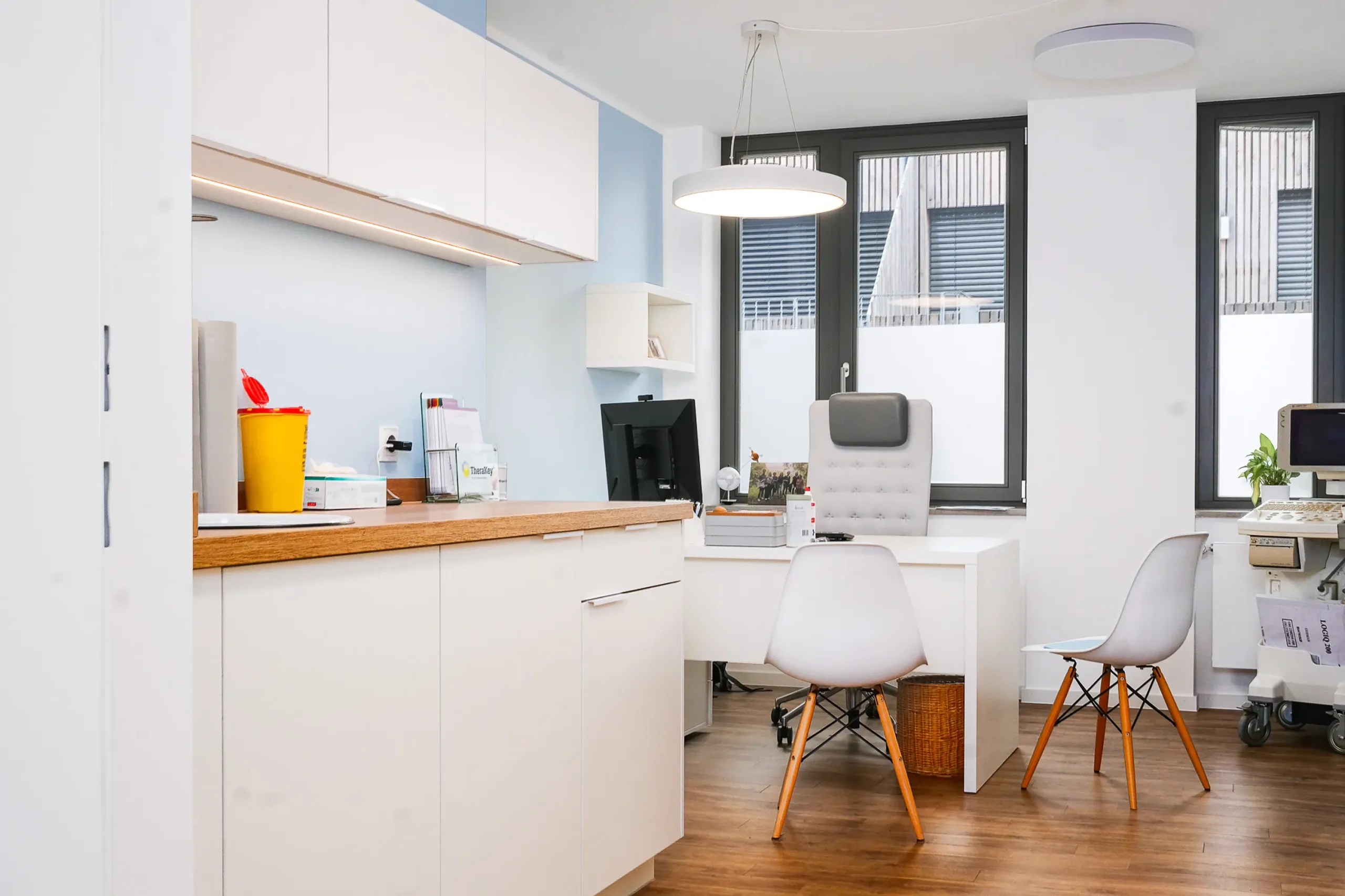 Modernes helles Arztpraxiszimmer mit Schreibtisch und Geräten Helles modernes Arztpraxiszimmer mit Schreibtisch, medizinischer Ausstattung und großen Fenstern.