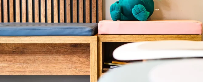 Moderner Wartebereich mit Holzbank, blauen und rosa Kissen und einem grünen Plüschkrokodil.