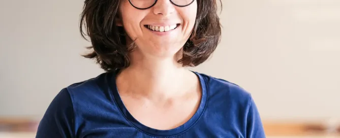 Frau mit Brille und dunkelblauem T-Shirt steht lächelnd in einer Praxisumgebung.
