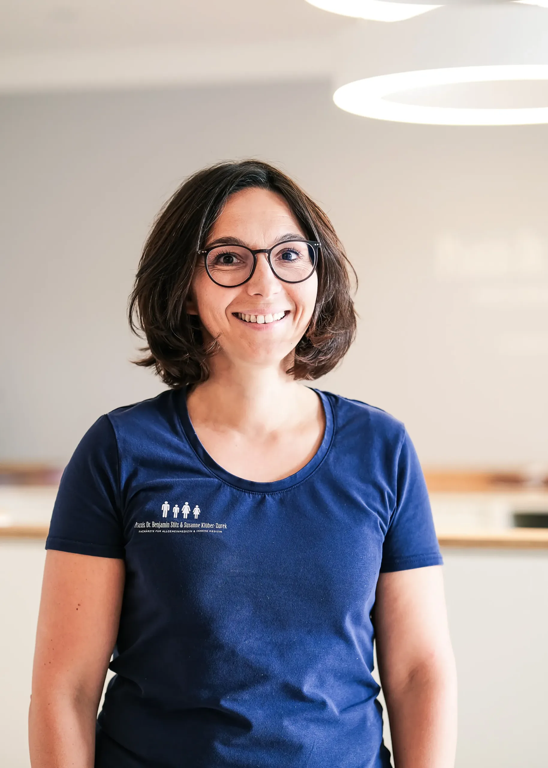 Freundlich lächelnde Frau mit Brille in Praxisumgebung Frau mit Brille und dunkelblauem T-Shirt steht lächelnd in einer Praxisumgebung.