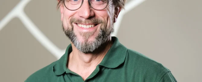 Markus Hirt mit Brille und grünem UWG-Poloshirt steht vor beige gemustertem Hintergrund.