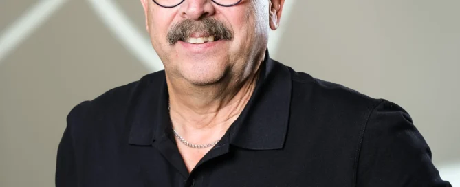Porträt von Martin Guggenberger mit Brille und schwarzem Polohemd vor grauem geometrischem Hintergrund.