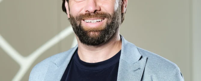 Porträt von Michael Dill mit Bart im grauen Blazer vor modern gemustertem Hintergrund.