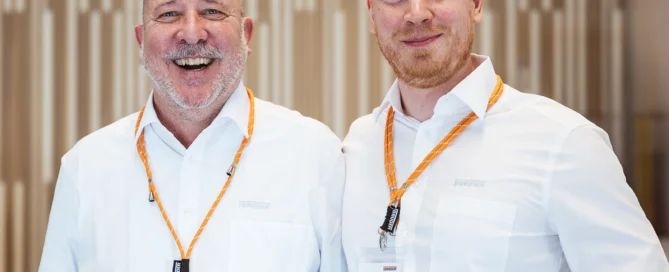 Zwei Männer bei Business-Event in Würzburg, smilend, in Hemden mit Lanyard und Namensschild vor modernem Hintergrund.