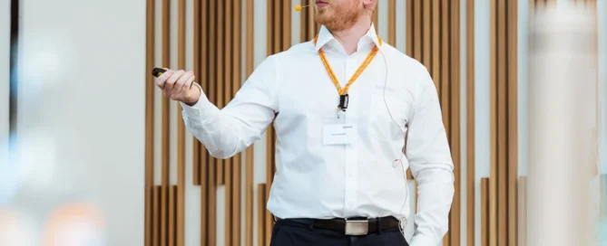 Mann hält Präsentation bei Business-Event in Würzburg, trägt weiße Hemd, dunkle Hose, Lanyard, gestikuliert.