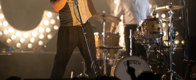 Eventfotografie eines Live Konzerts in Frankfurt: Sänger Dick Brave zeigt Peace-Zeichen, hinter ihm leuchtende Buchstaben, Publikum im Dunkeln