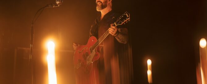 Chris Vega mit roter Gitarre, im warm beleuchteten Vintage-Studio, vor Mikrofon und dunklem Hintergrund.