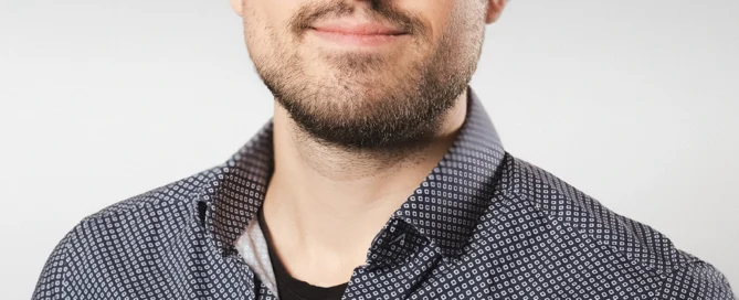 Porträt eines jungen Mannes in Businesskleidung mit Brille vor grauem Hintergrund, professionell und ruhig.