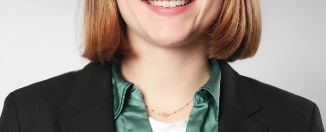 Professionelles Porträt einer Frau in Businesskleidung, mit Brille, Lächeln und neutralem grauem Hintergrund.