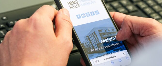 Person hält Smartphone mit Immobilienbild und Text, am Schreibtisch mit Tastatur und Wasser im Büro.