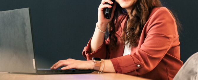 Frau im Business-Outfit am Schreibtisch, telefonierend mit Laptop vor Neon-Schriftzug IMMO HELLER in Würzburg.