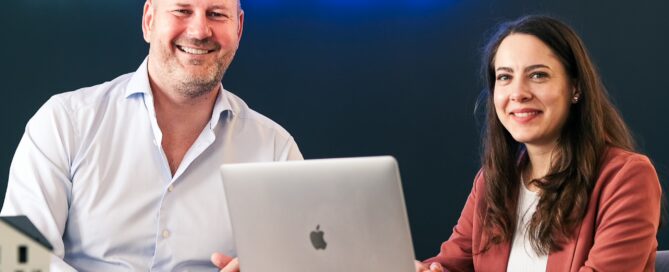 Johannes Heller und Mitarbeiterin beim Meeting in einem modernen Büro mit Laptop, Neon-Schriftzug und Häuser-Modell.