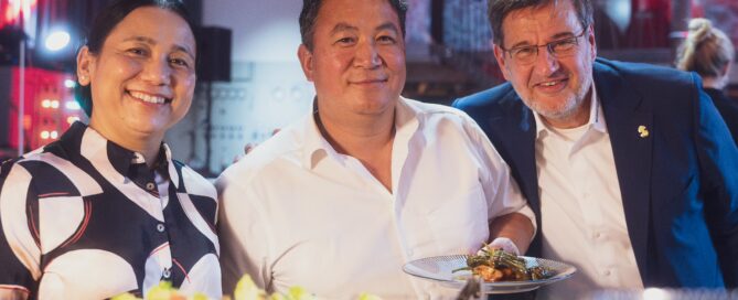 Drei Menschen bei einem festlichen Abendessen in Würzburg, lachen, mit Essen und warmem Licht im Hintergrund.