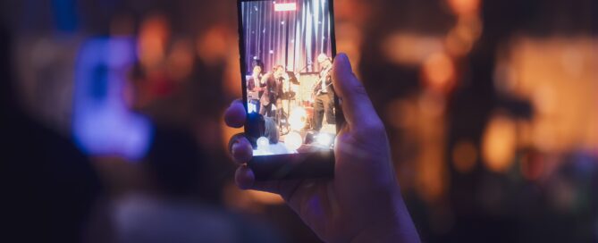 Person filmt Live-Musikperformance mit Smartphone, fokussiert auf das Display, bei gedämpften Bühnenlichteffekten.