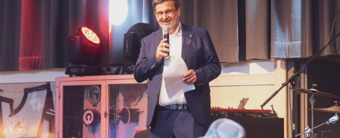Simon Wagner hält Rede auf  Jubiläumsveranstaltung 70 Jahre Wagner Sicherheitstechnik AG in Würzburg, umgeben von Publikum und Bühnenausstattung.